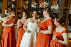 Bridal party wearing vibrant orange dresses and white wedding gown gather for candid moment at wedding reception.