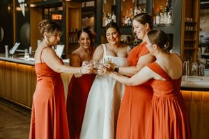 A wedding celebration at a bar with attendees wearing orange bridesmaid dresses toasting with champagne glasses.