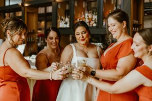 Wedding party wearing white and orange dresses toasting with glasses in a bar setting with warm lighting.