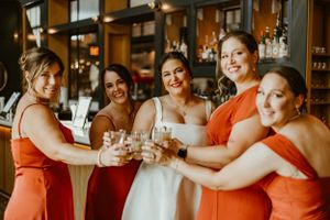 Bridal party wearing orange dresses toasts with glasses in a warmly lit venue interior.