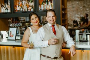 A couple stands together at a bar counter with liquor bottles displayed on shelves behind them.