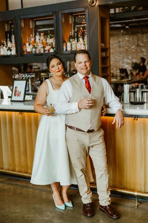 Couple in elegant attire posing at a bar with exposed brick wall and liquor display.
