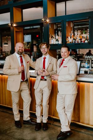 Three groomsmen in matching beige suits and red ties pose together at a stylish bar counter.
