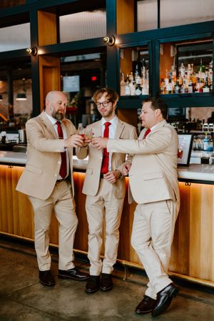 Three groomsmen in beige suits sharing drinks at a wooden bar counter.