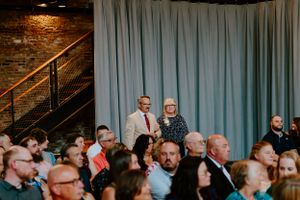 An audience sits attentively in rows at an indoor venue with exposed brick walls and teal curtains in the background.