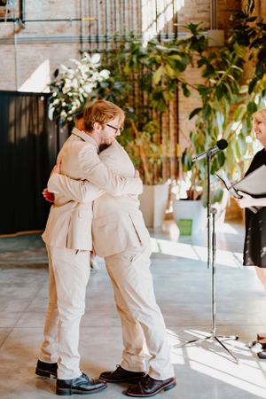 Two people in matching beige outfits embrace during an indoor wedding ceremony.
