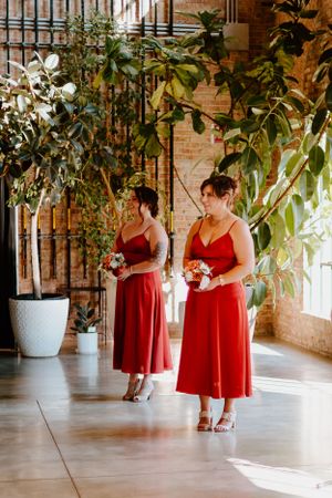 Two bridesmaids in red dresses standing near potted plants in a sunlit brick interior.