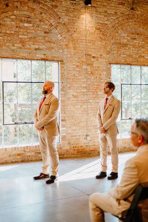Two groomsmen in tan suits standing in a sunlit brick room with large windows.