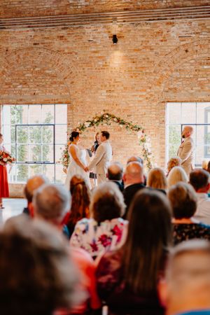 Wedding ceremony in a rustic brick venue with floral arch and guests watching the ceremony.