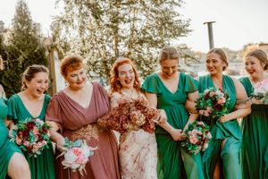 A bridal party in green and pink dresses laugh together while holding bouquets outdoors.