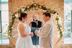 Wedding ceremony under floral arch with brick backdrop in natural light.