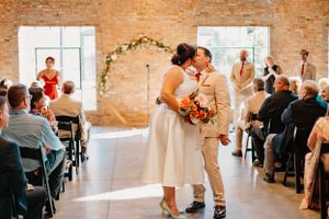 A couple celebrating their first kiss as newlyweds in a rustic brick venue with natural light streaming through windows.