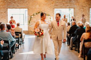 A wedding couple walks down the aisle in a rustic brick venue under a floral arch.