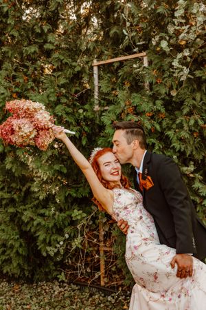 A joyful wedding couple poses against greenery with the bride holding a pink bouquet overhead in a candid moment.