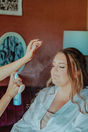 A stylist sprays hairspray while preparing someone in a white robe during a beauty session.
