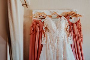 A white lace wedding dress hangs between coral bridesmaid dresses on a clothing rack in soft natural light.