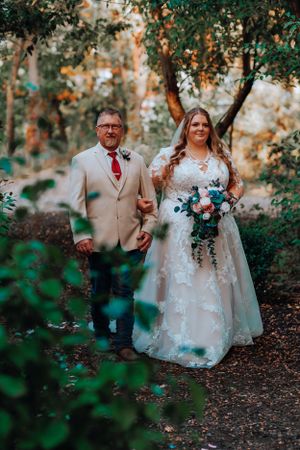 A couple in wedding attire stands together on a woodland path surrounded by green foliage during golden hour.