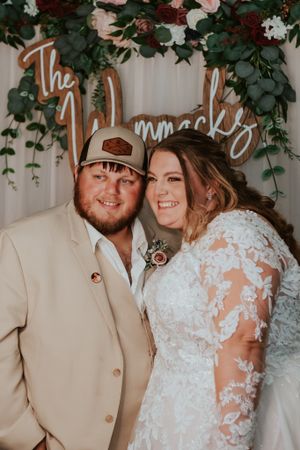 A bride in a lace dress and groom in tan suit pose under wedding decor with greenery and signage.