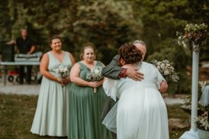 Two women in wedding attire embrace during an outdoor ceremony while bridesmaids in sage green dresses look on.