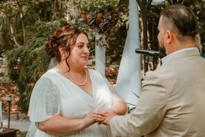 A couple exchanges rings during an outdoor wedding ceremony against a backdrop of trees and white pillars.
