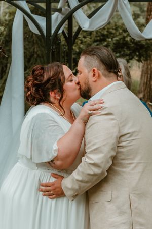 Newlyweds share their first kiss under white fabric draped arbor at an outdoor wedding ceremony.