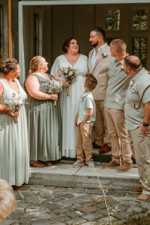 Wedding party in light beige and sage green attire gather on stone steps for outdoor photos.