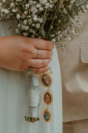 Close-up of hands holding white baby's breath bouquet with vintage locket charms and delicate ring detail.