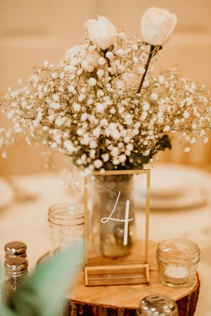 Rustic wedding centerpiece featuring baby's breath and white roses in a mason jar with table number 4 on wood slice.