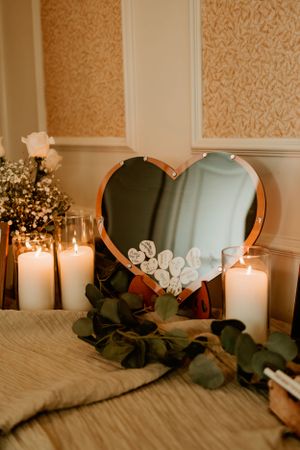 Heart-shaped mirror adorned with white flowers sits among lit candles and eucalyptus leaves on a textured surface.
