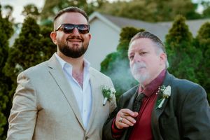 Two wedding guests in formal attire pose together outdoors while one exhales cigar smoke.