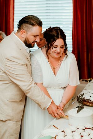 A couple in wedding attire cut their cake together at their wedding reception, dressed in beige and white.
