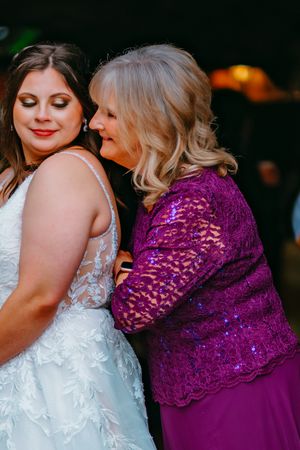 Two women share a tender moment at a wedding, with one wearing a white lace gown and the other in a vibrant purple dress.