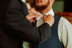 Someone adjusts a light gray bowtie on a groom wearing a navy vest and white dress shirt.