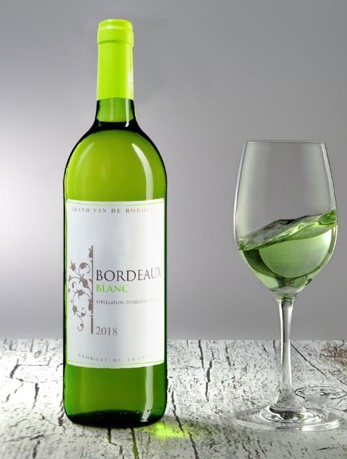 A bottle of Bordeaux white wine and a glass of white wine on a rustic white wooden surface.