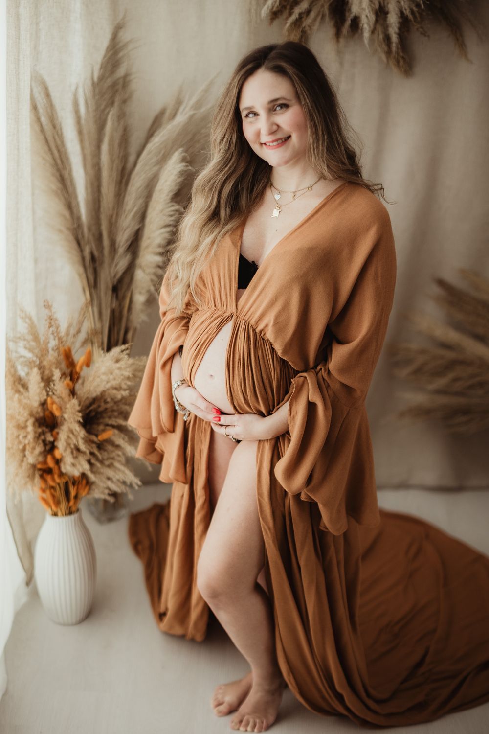 Maternity photo series in warm earth tones featuring flowing rust-colored dress and dried floral arrangements in vases.