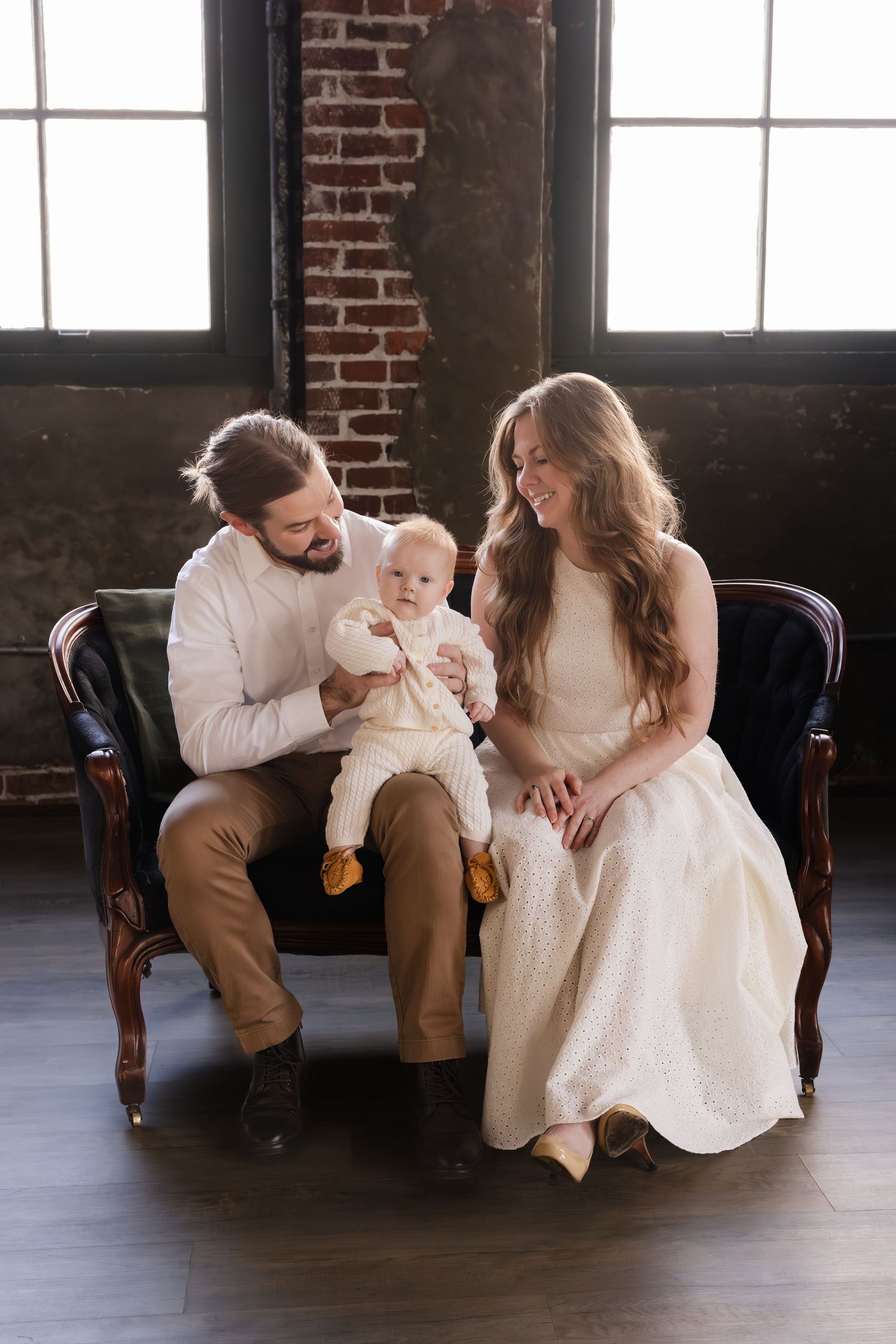 Young family sits together on antique sofa in rustic brick loft with natural window light.