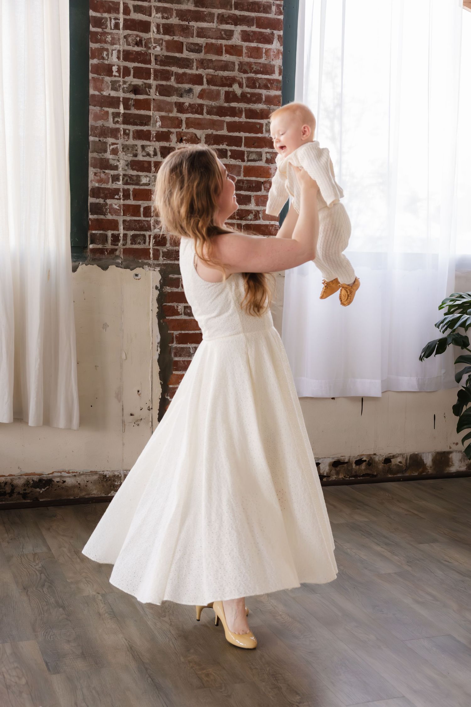 A person in a white dress dances while holding up a baby in white against a rustic brick wall interior.