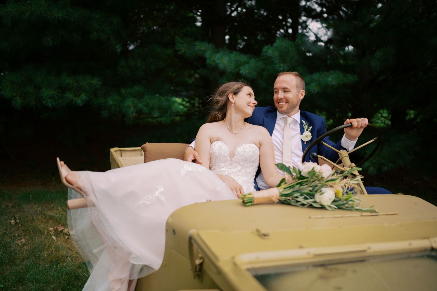 A couple sits together at a rustic outdoor table, sharing a tender moment in formal wedding attire amid lush greenery.
