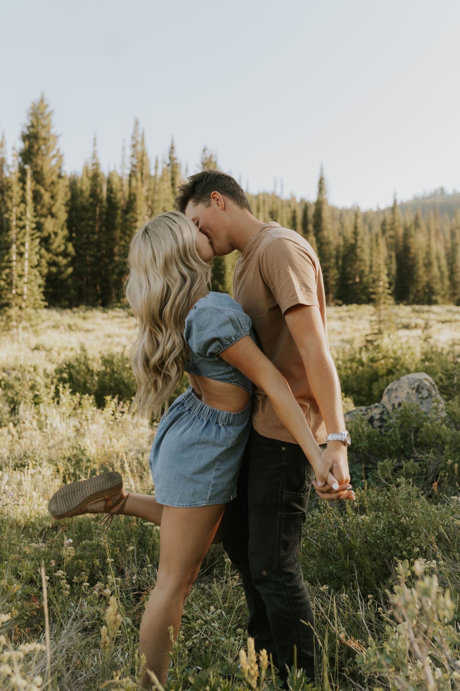 A romantic couple embraces in a serene mountain meadow surrounded by evergreen trees on a sunny afternoon.