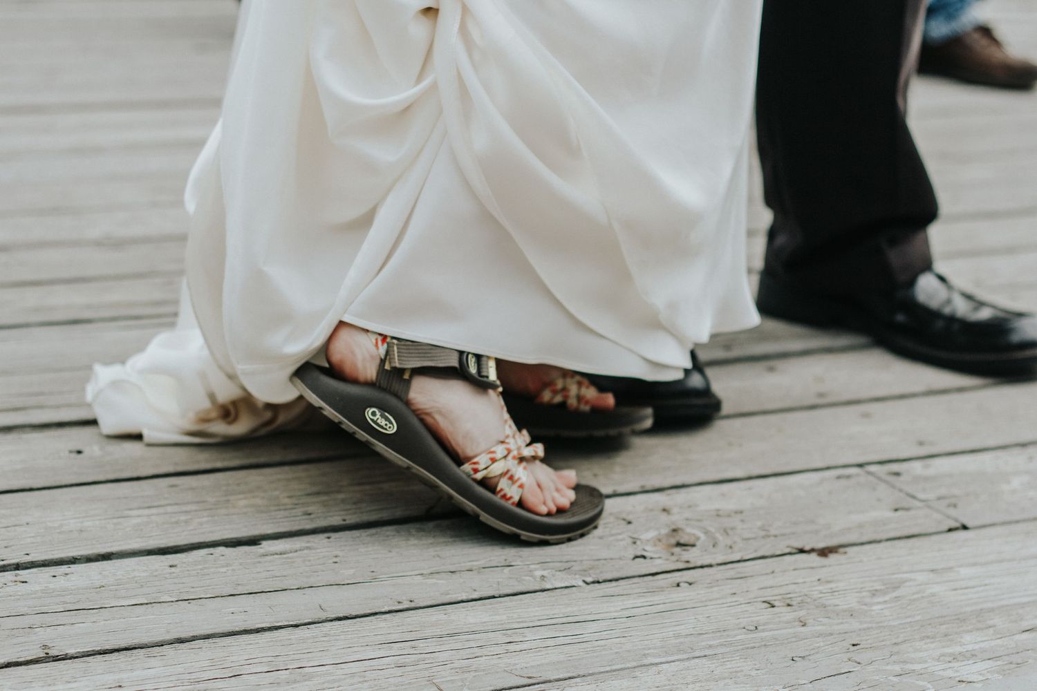 Close-up of sandaled feet and flowing white dress hem on a wooden deck.
