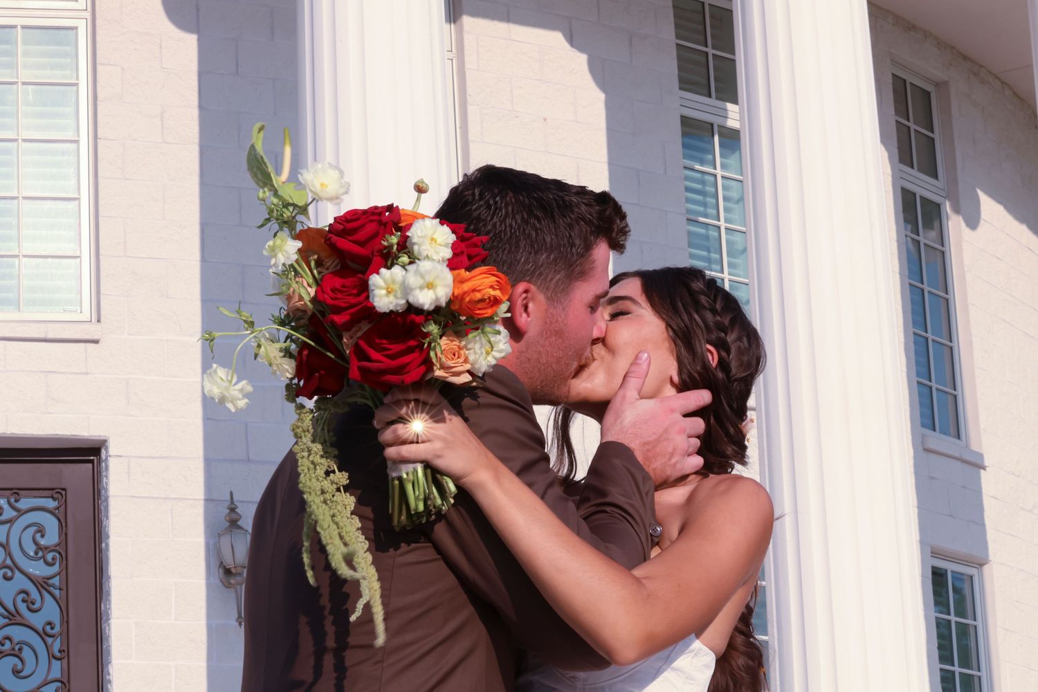 A couple shares a romantic kiss on their wedding day while holding a vibrant red and white bouquet.