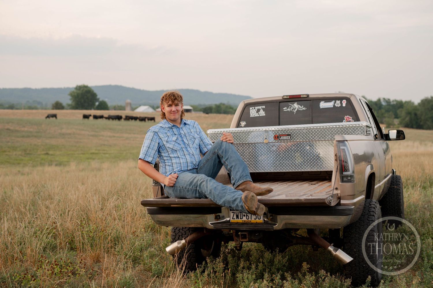 A person sits casually on the tailgate of a silver pickup truck in a rural field.