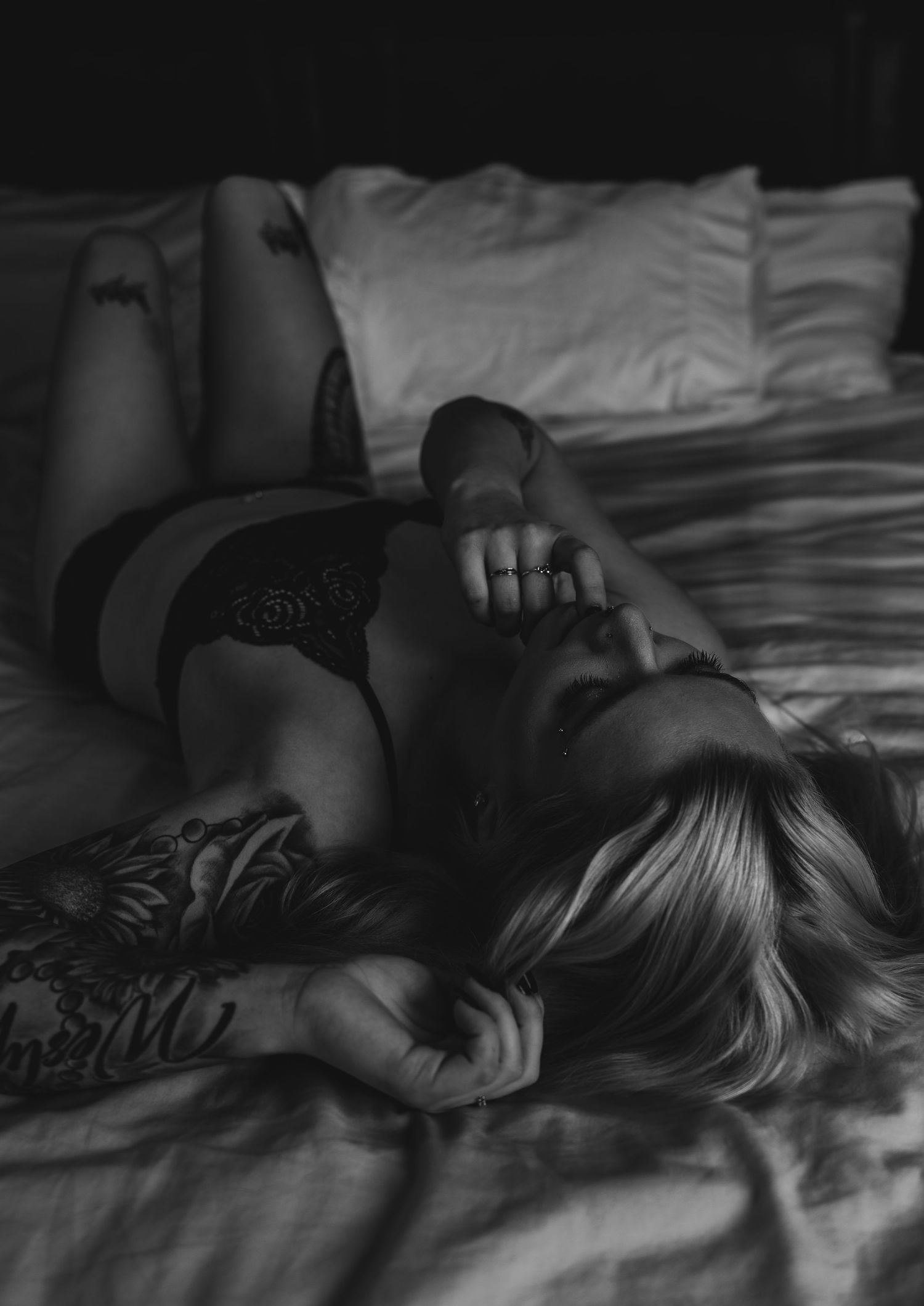 Moody black and white boudoir photo of a woman with tattoos lying on a bed.