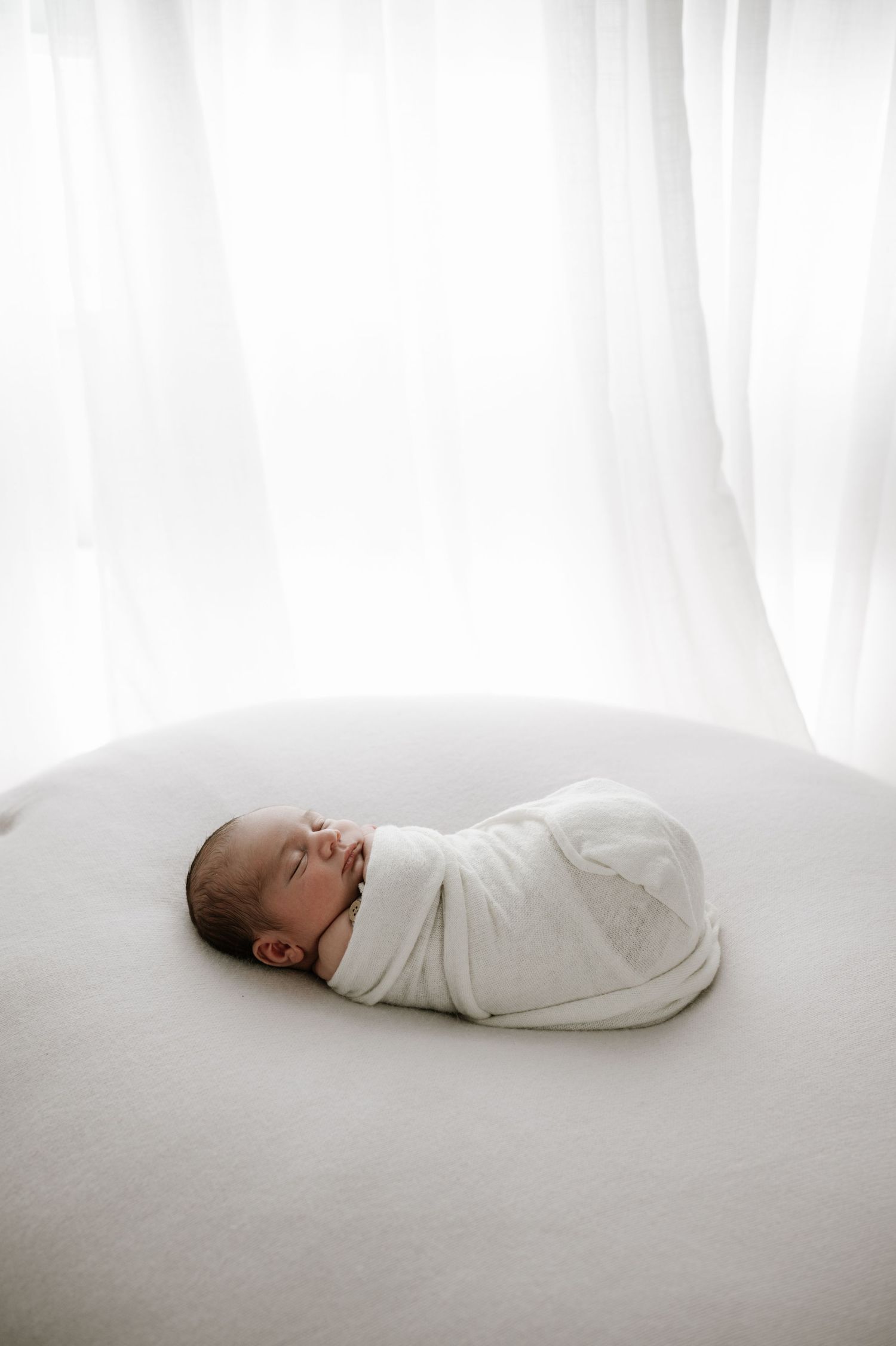 A newborn baby wrapped in white swaddle cloth sleeps peacefully on a white surface against a light background.