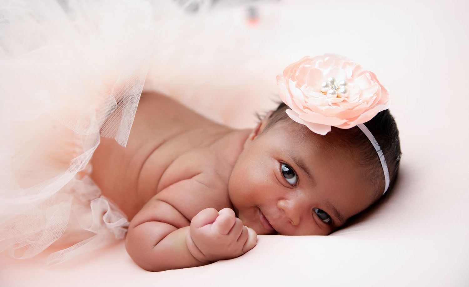 Baby wearing pink flower headband lies on soft tulle fabric in portrait pose.