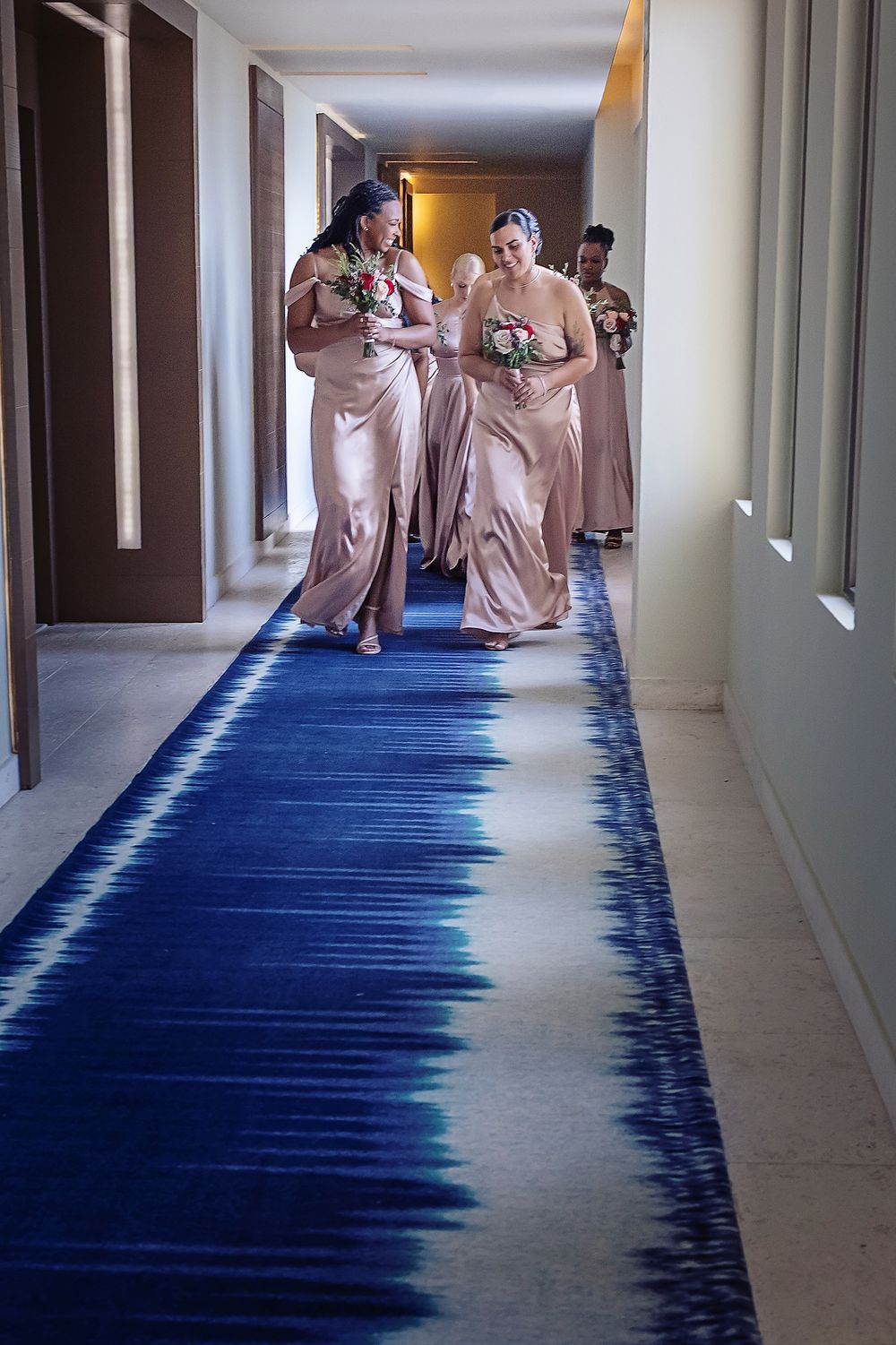 Bridesmaids in blush pink dresses walk down a hallway with a striking blue and white tie-dye patterned carpet.