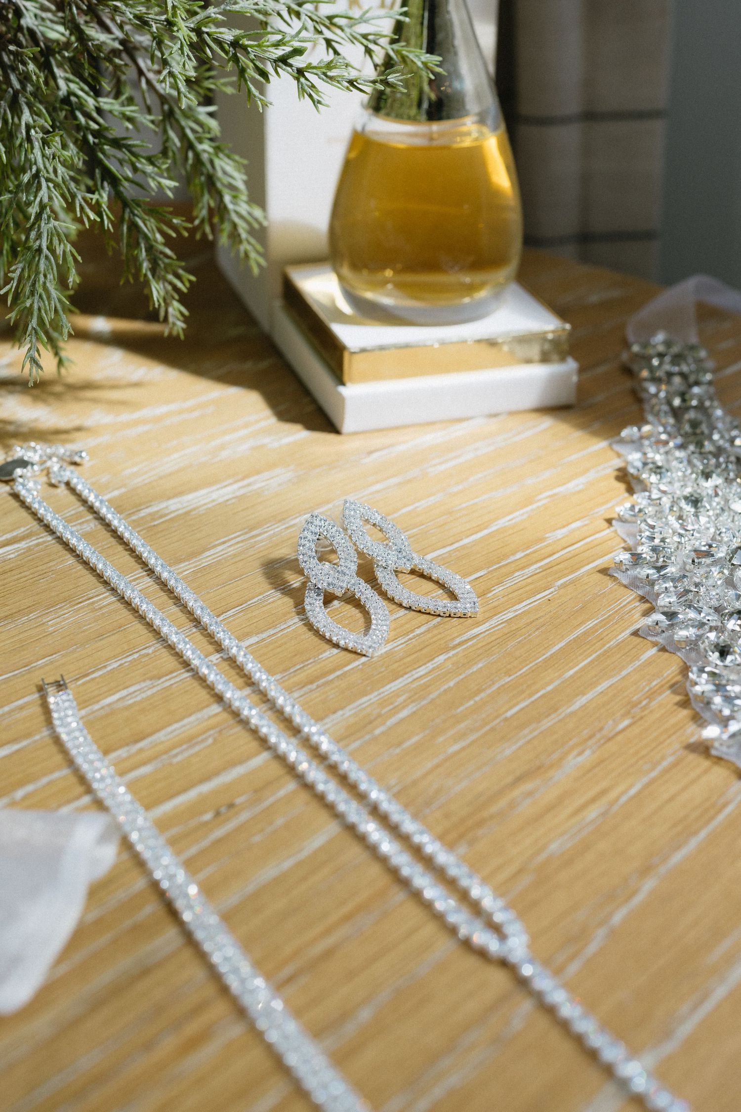 Pearl necklace and diamond ring arranged on wooden surface with olive oil bottle and greenery.