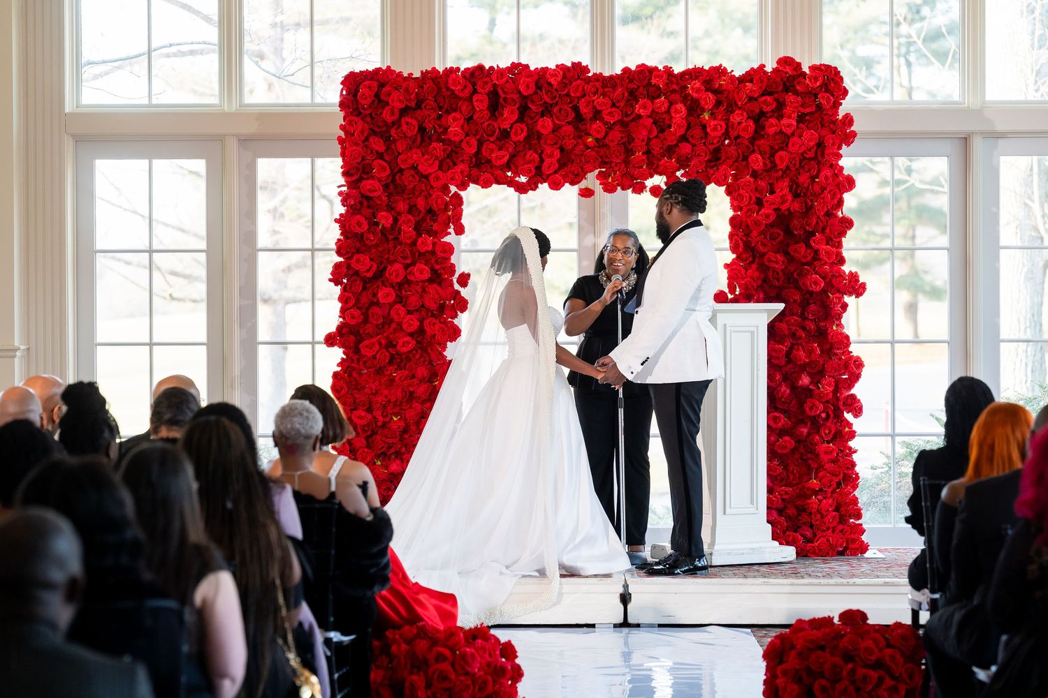 Wedding ceremony with bride and groom at red rose floral arch altar in elegant white venue. Deerfield Golf Club Newark De