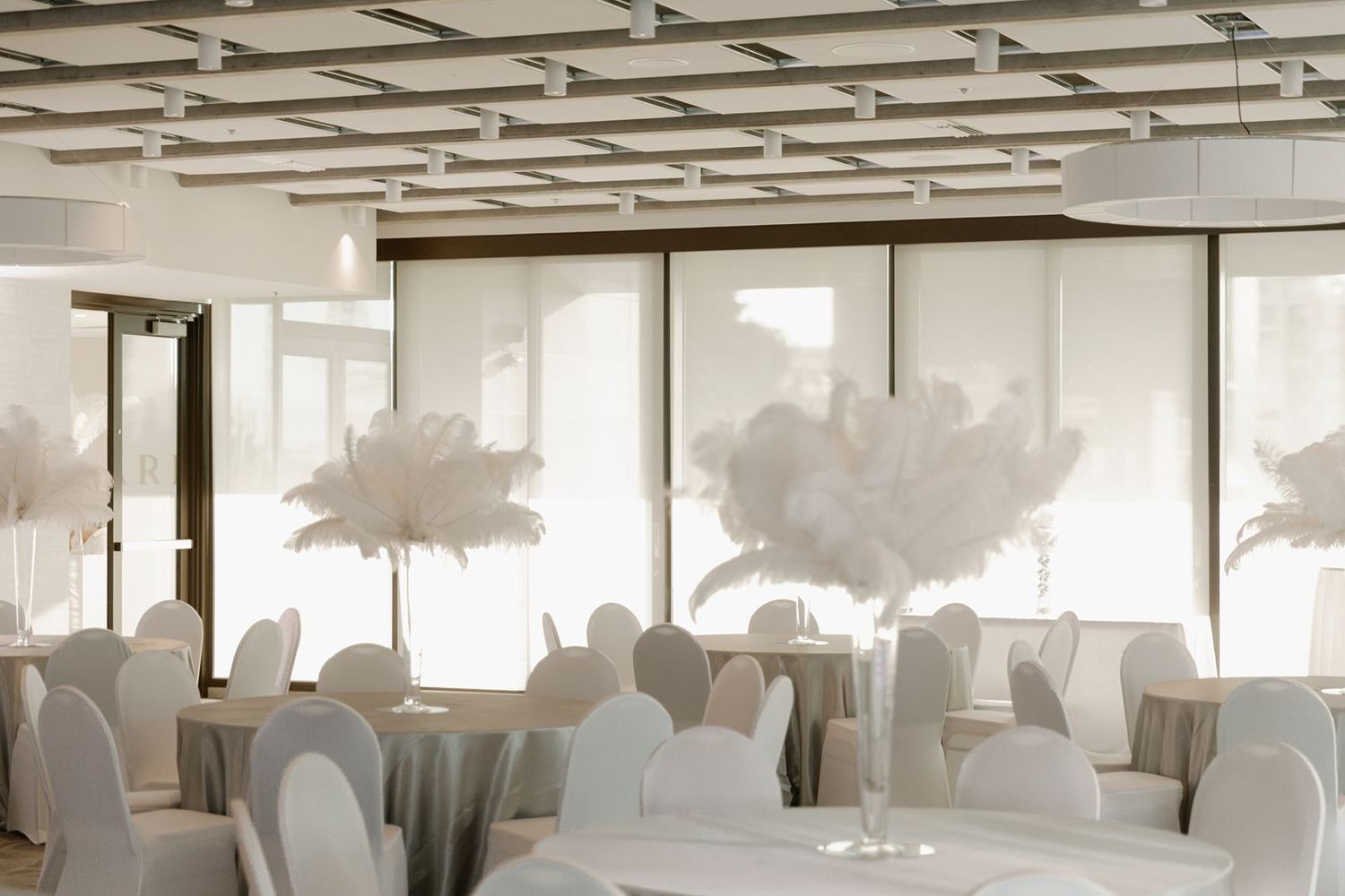 Bright wedding reception hall with white feather centerpieces and white chair covers.