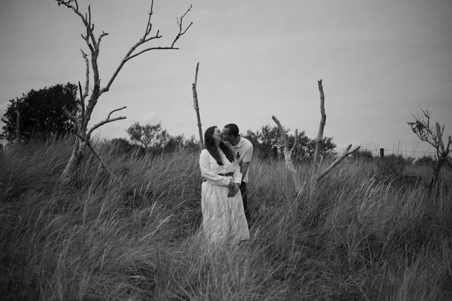Black and white photo of a couple kissing in tall grass with bare trees in a romantic outdoor setting.